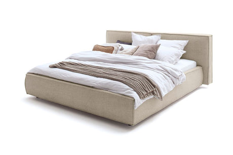Ein beige gepolstertes Bett mit weißer Bettwäsche, Kissen und einer braunen Überwurfdecke vor weißem Hintergrund.